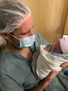 Photo of a dad wearing a hair net, surgical mask, and blue scrubs holding a newborn baby.